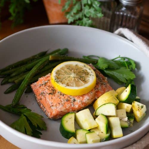 piece of fish / salmon on a grey plate with green beans and zuchini and summer squash. Lemon is on top of the fish. background there are salt and pepper shakers and a plant