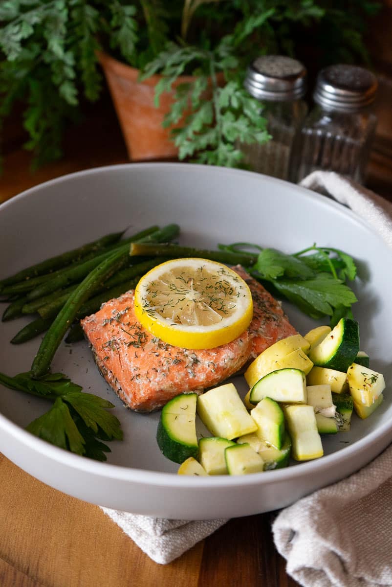 piece of fish / salmon on a grey plate with green beans and zuchini and summer squash. Lemon is on top of the fish. background there are salt and pepper shakers and a plant