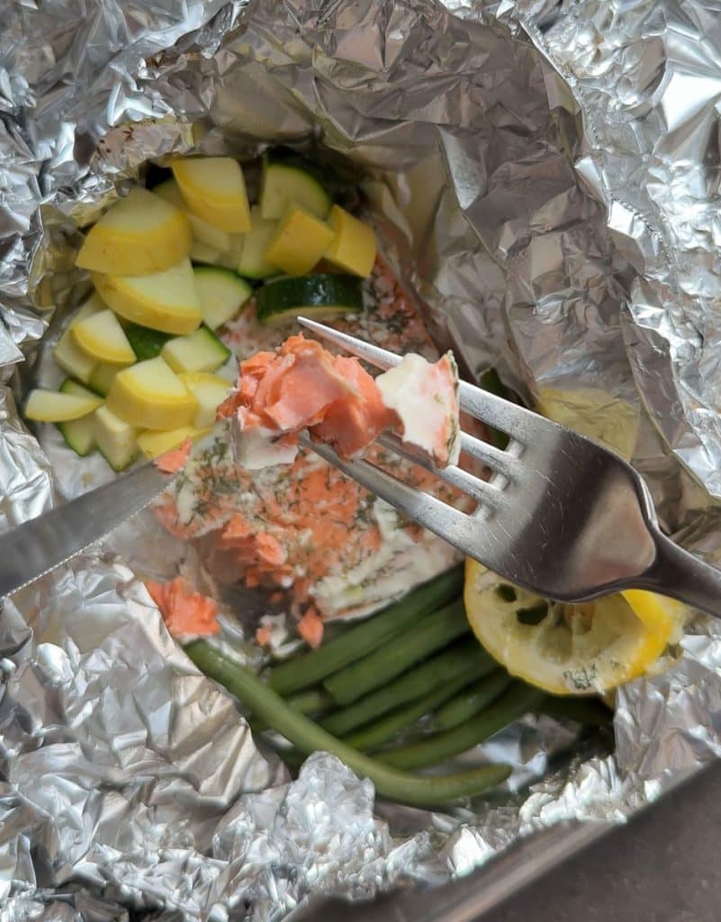 a fork with some fish on it with the baked packet of fish and vegetables are in the background