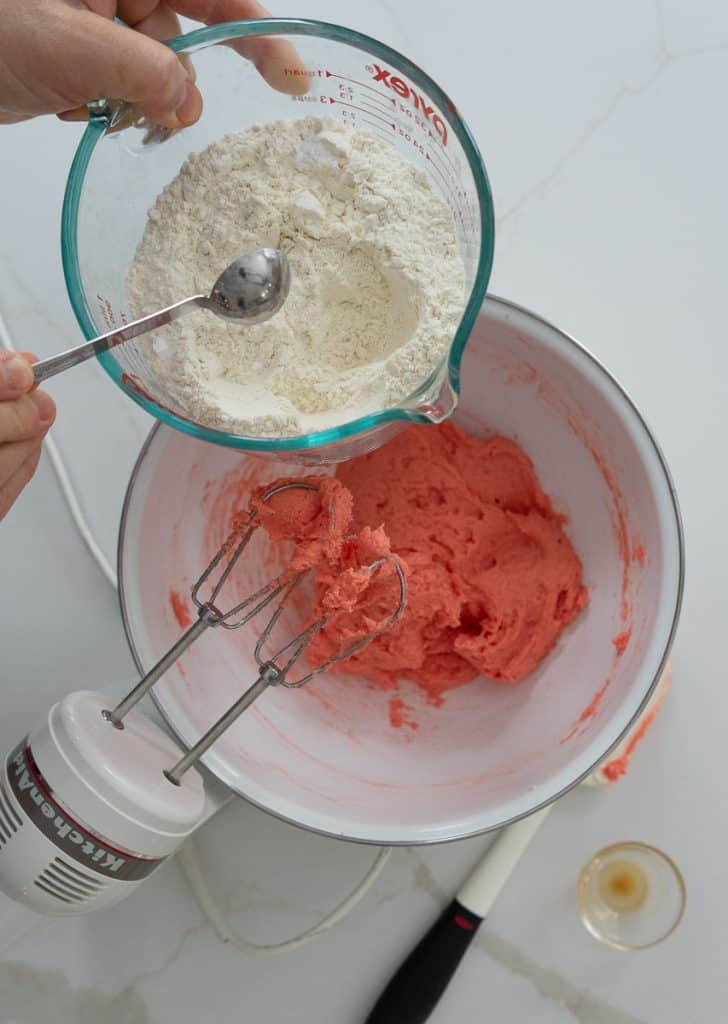 combining dry ingredients in a measuring cup and adding to the wet ingredients,  mixer is in the lower right corner, dry ingredients are being added to the bowl from the top