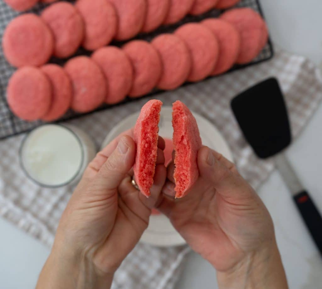 pink jello cookies on a cooling rack with a plate of cookies and a glass of milk there is a spatula in the right lower corner two hands are breaking a cookie in half over the plate of cookies