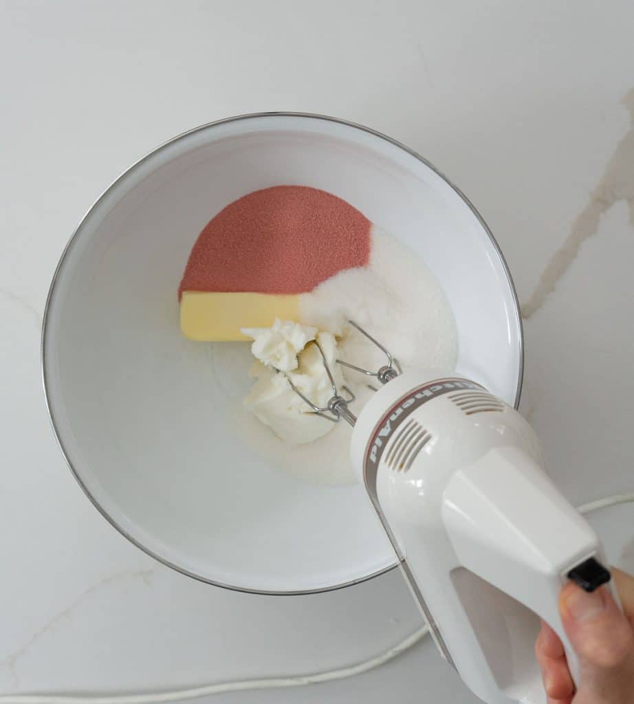 butter, shortening, and jello in a white bowl with a hand holding a mixer in the lower right hand corner ready to mis