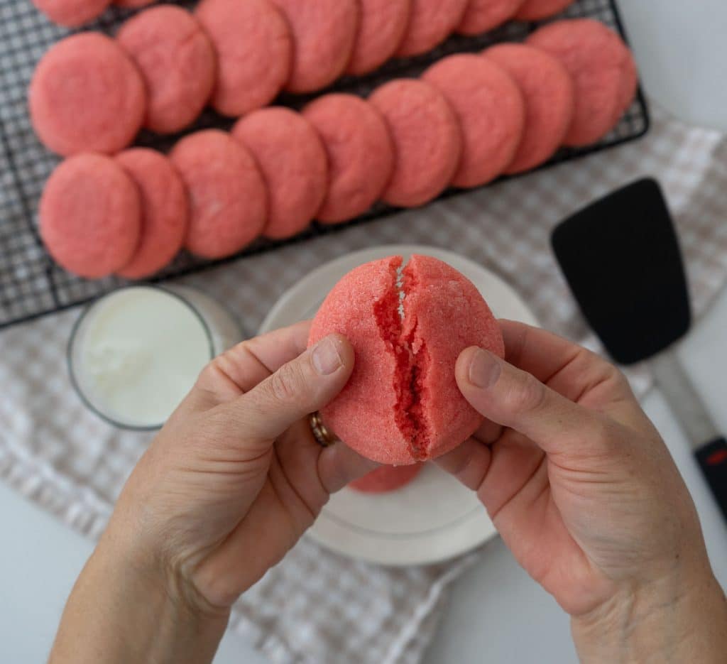 pink jello cookies on a cooling rack with a plate of cookies and a glass of milk there is a spatula in the right lower corner two hands are breaking a cookie in half over the plate of cookies