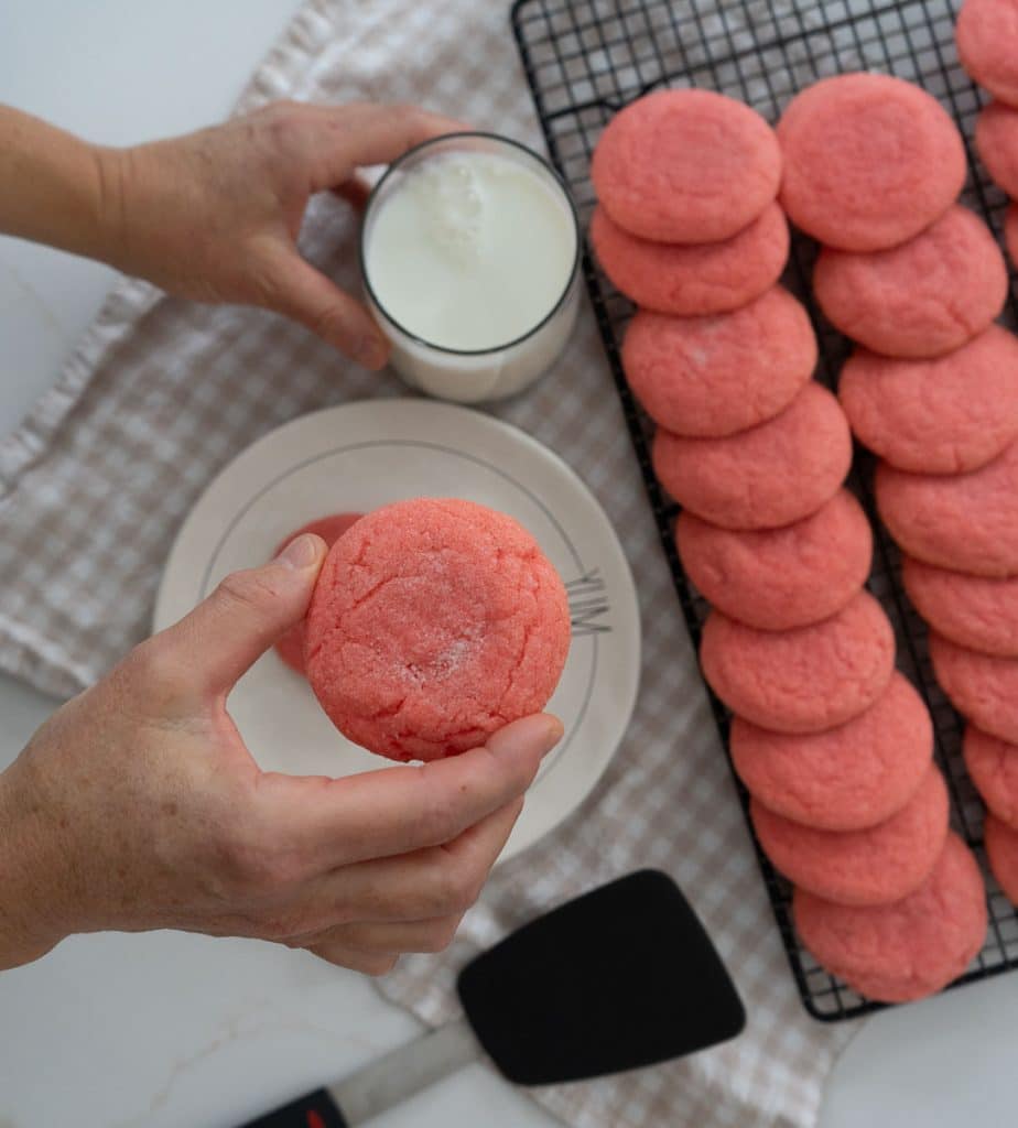 rack of jello cookies cooling with a plate of cookies in front of it, a hand is taking a cookie from the plate and another hand is coming in from the left and lifting a glass of milk