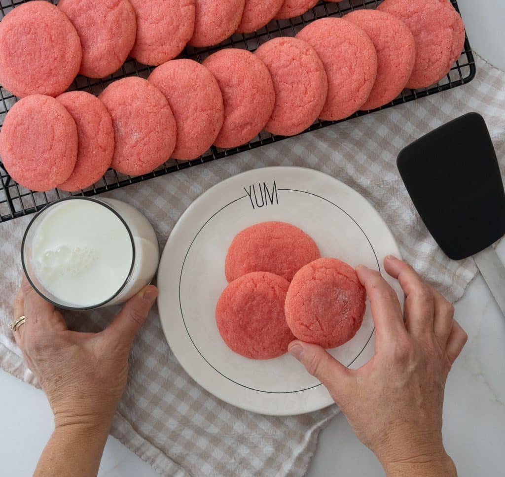 rack of jello cookies cooling with a plate of cookies in front of it, a hand is taking a cookie from the plate and another hand is coming in from the left and lifting a glass of milk