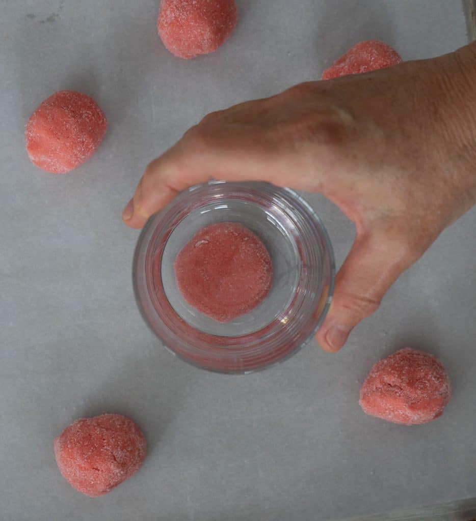 cookie dough on parchment lined baking sheet a hand is holding a juice glass and pressing into the cookie dough to flatten