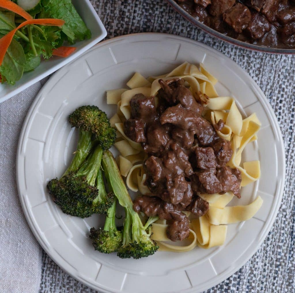plate of no peek beef tips on top of noodles with broccoli red pot in the background