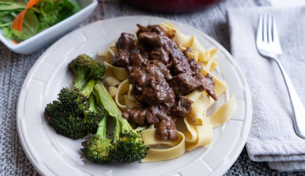 plate of no peek beef tips on top of noodles with broccoli red pot in the background