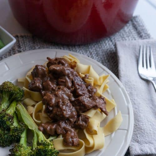 plate of no peek beef tips on wide egg noodles with broccoli red pot of no peek beef tips in the background