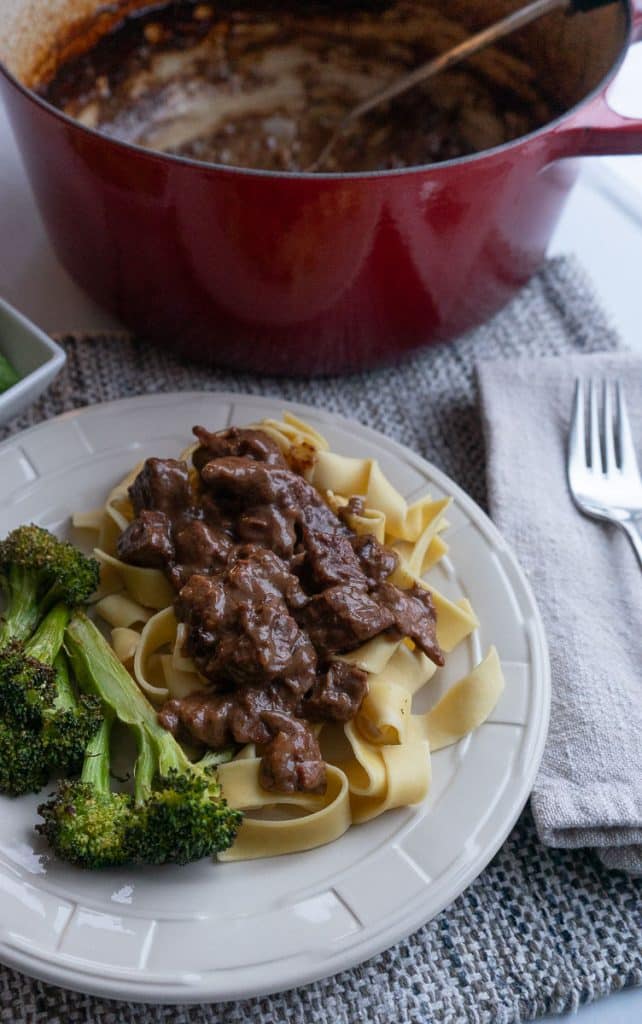 plate of no peek beef tips on wide egg noodles with broccoli red pot of no peek beef tips in the background