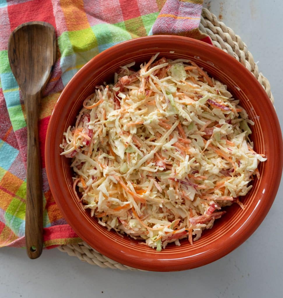 ginger coleslaw in a rust colored bowl that is sitting on a colorful kitchen towel, there is a wooden spoon laying to the left of the bowl