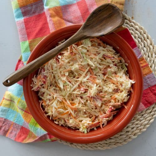 ginger coleslaw in a rust colored bowl that is sitting on a colorful kitchen towel, there is a wooden spoon laying across the bowl