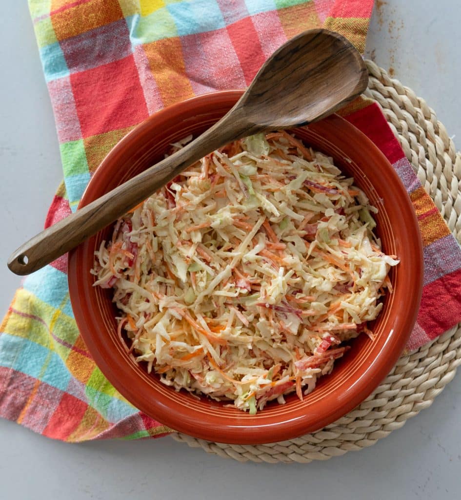 ginger coleslaw in a rust colored bowl that is sitting on a colorful kitchen towel, there is a wooden spoon laying across the bowl