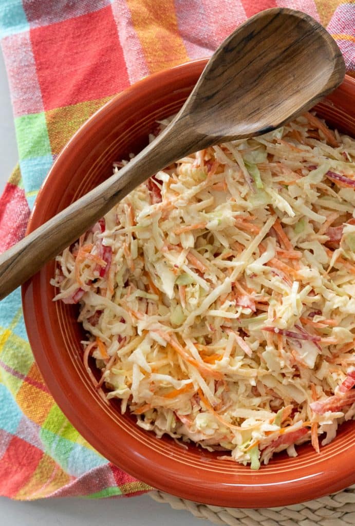 ginger coleslaw in a rust colored bowl that is sitting on a colorful kitchen towel, there is a wooden spoon laying across the bowl