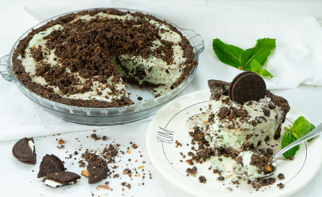 whole ice cream pie with one piece cut out is on the left side of the photo, the slice is on a white plate on the right side with a sprig of mint cookies are crumbles around the plate