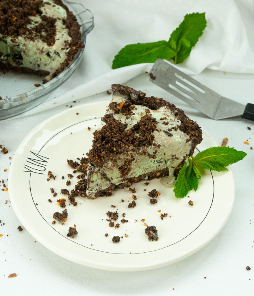 Slice of mint chocolate chip ice cream pie on a plate with whole pie in the back ground