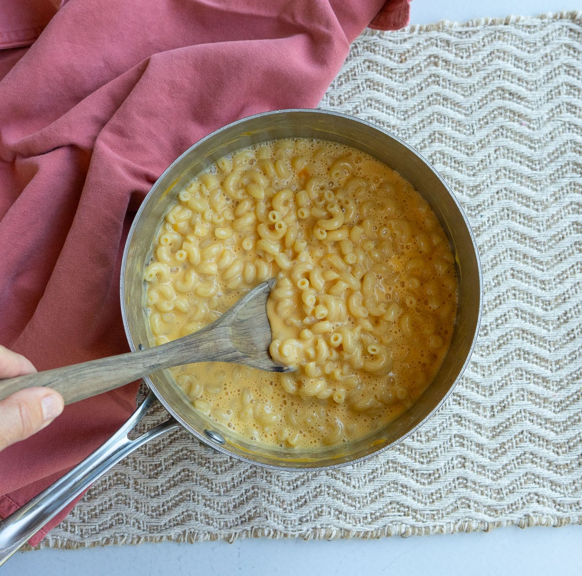 pan on a rattan mat with stovetop mac n cheese in the pan with a wooden spoon being stirred by a hand there is a rust colored cloth in the upper left coner