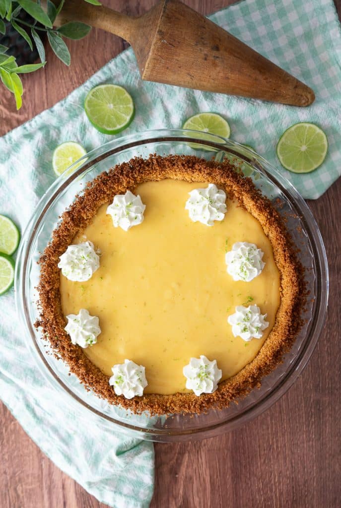 picture of the whole pie decorated with whipping cream and lime zest on a wood countertop with a green and white gingham cloth underneath the pie