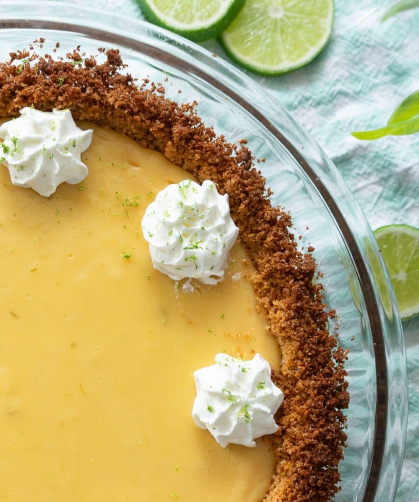 picture of a quarter of key lime pie in a pie plate on a green and white gingham cloth. with lime slices laying around the pie plate