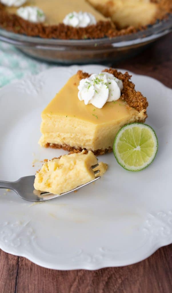 piece of key lime pie with graham cracker crust sitting on a white plate there is a fork in the forefront of the photo with a bite of key lime pie on a fork setting on the plate