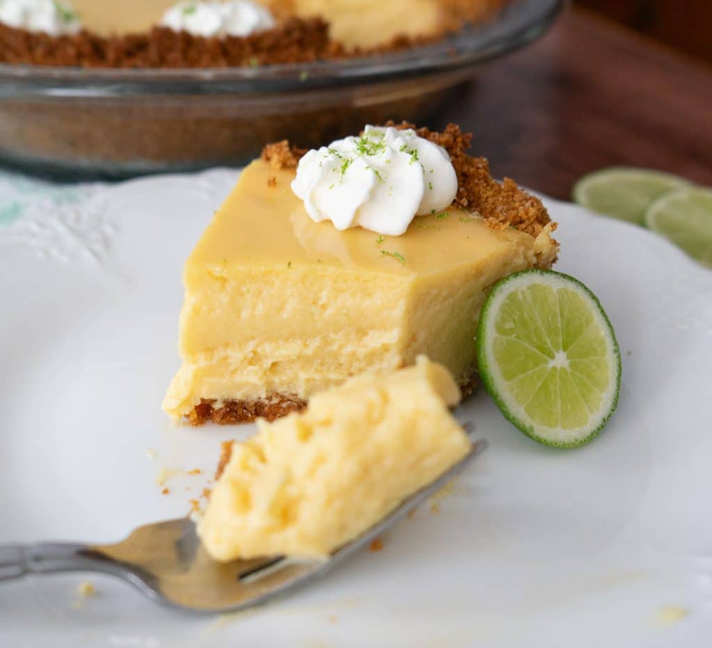 piece of key lime pie with graham cracker crust sitting on a white plate there is a fork in the forefront of the photo with a bite of key lime pie on it the whole pie is in the background