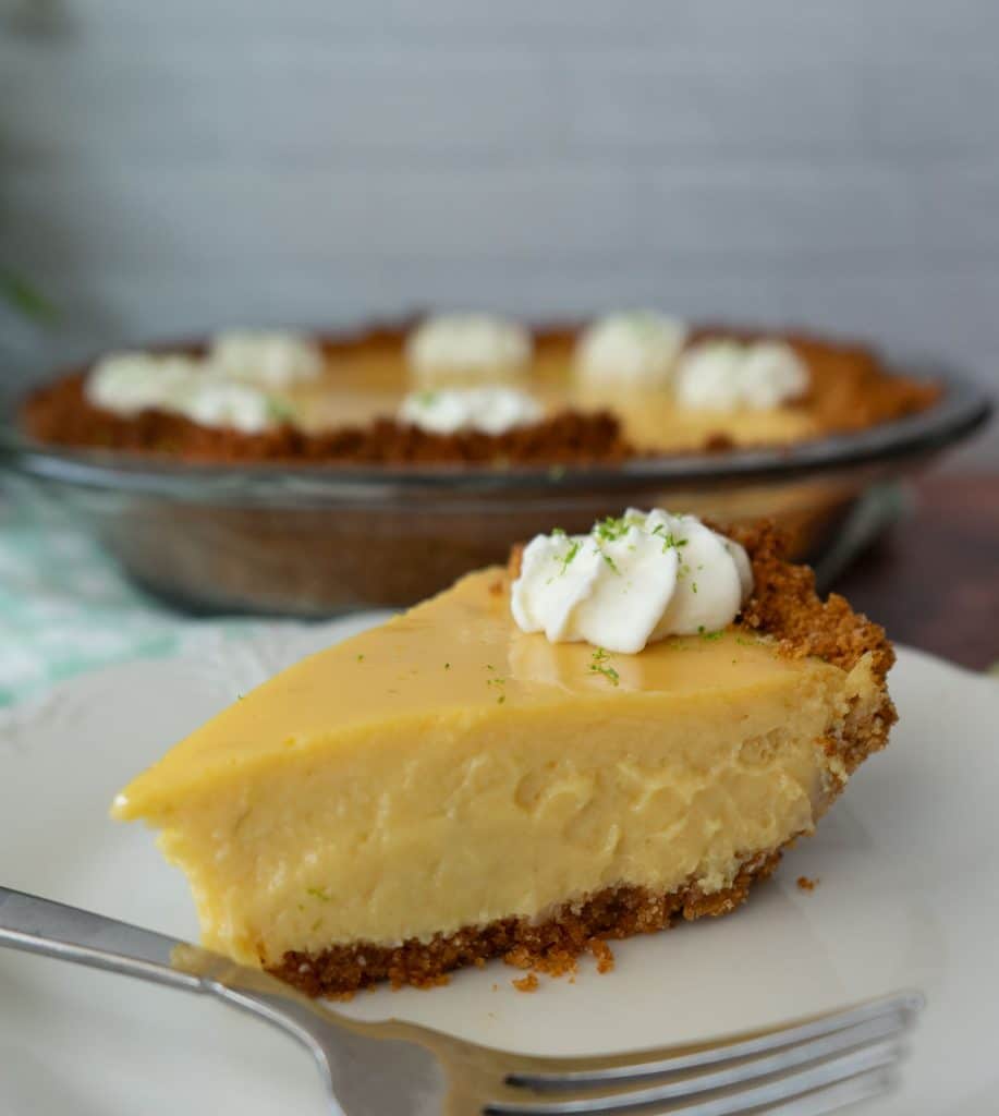 one piece of pie on a white plate with a fork on the plate the whole pie with a piece taken out is in the background of the photo
