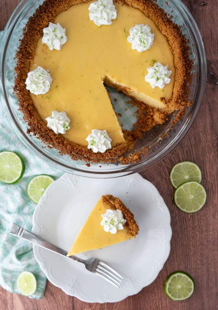 top view of whole key lime pie with one slice taken out and a piece of pie on a white plate with a fork on the plate there are slices of limes laying around 