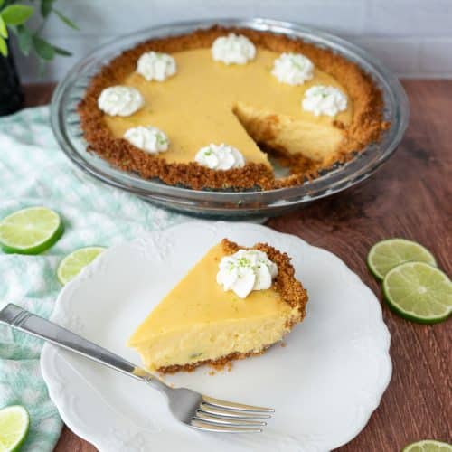 one piece of key lime pie on a white china plate with a fork on the plate next to the piece of pie the whole pie is in the background of the photo picture taken on a wood table top
