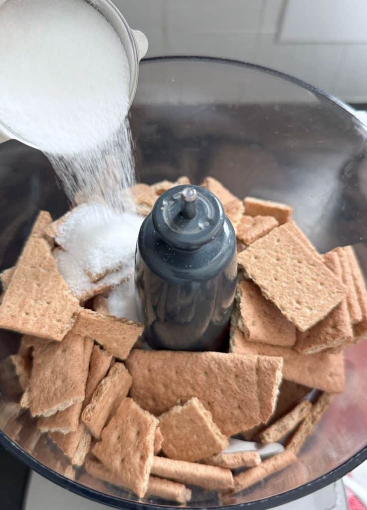 graham crackers in the bowl of a food processor sugar is being poured in to the bowl