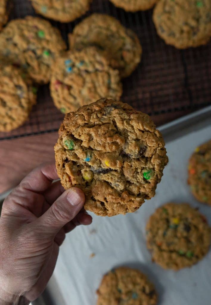 hand holding a monster cookie over the pan of cookies and cookies cooling