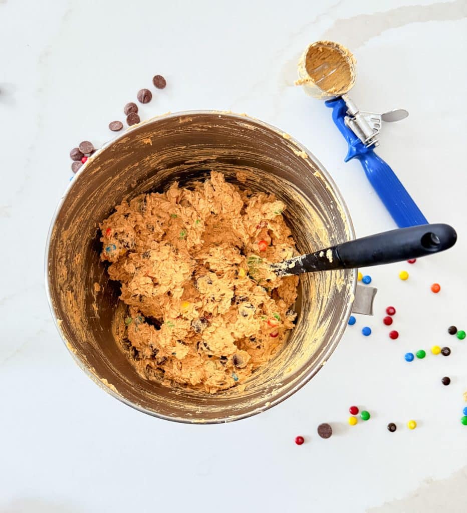 cookie dough in stainless steel bowl on white counter with m &ms sprinkled around on countertop