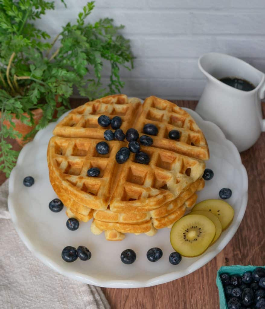 a plate stacked with waffles that have blueberries and kiwi on them with syrup in the background in a white glass container
