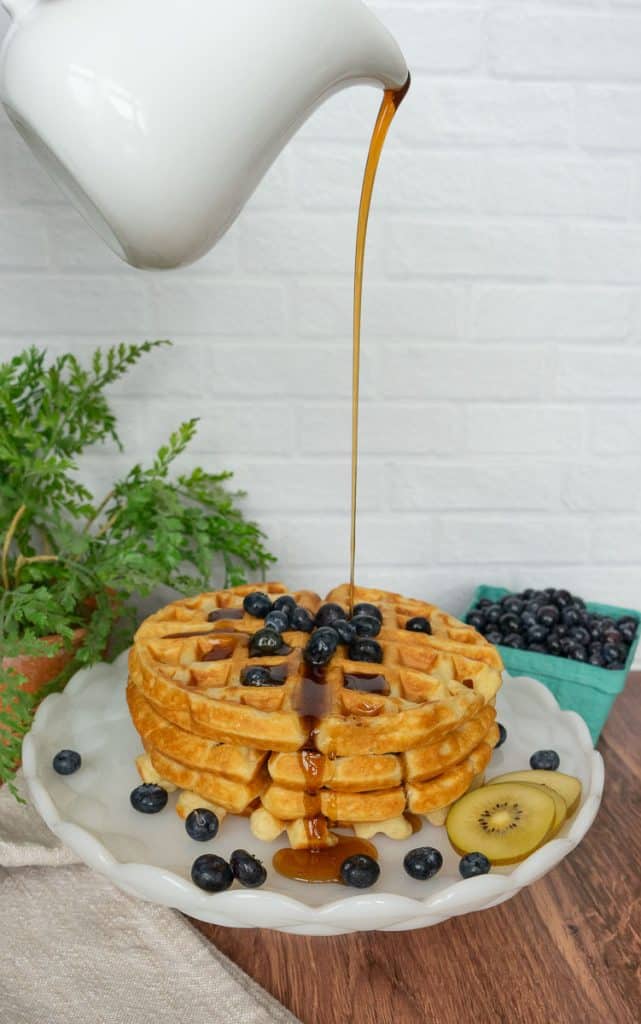 pouring syrup over a stack of waffles on a plate the waffles have blueberries on them and kiwis on the side with a small container of blueberries in the background and a plant on one side in the back ground