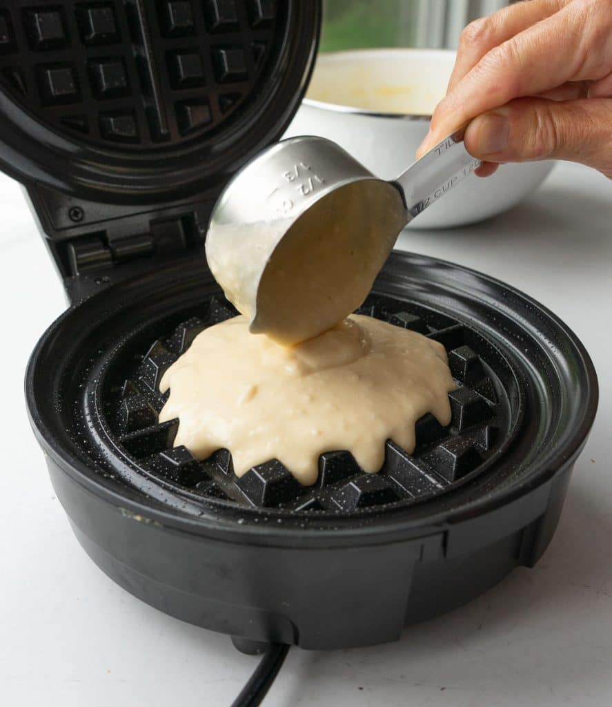 pouring batter in to a preheated waffle iron with a silver measuring cup