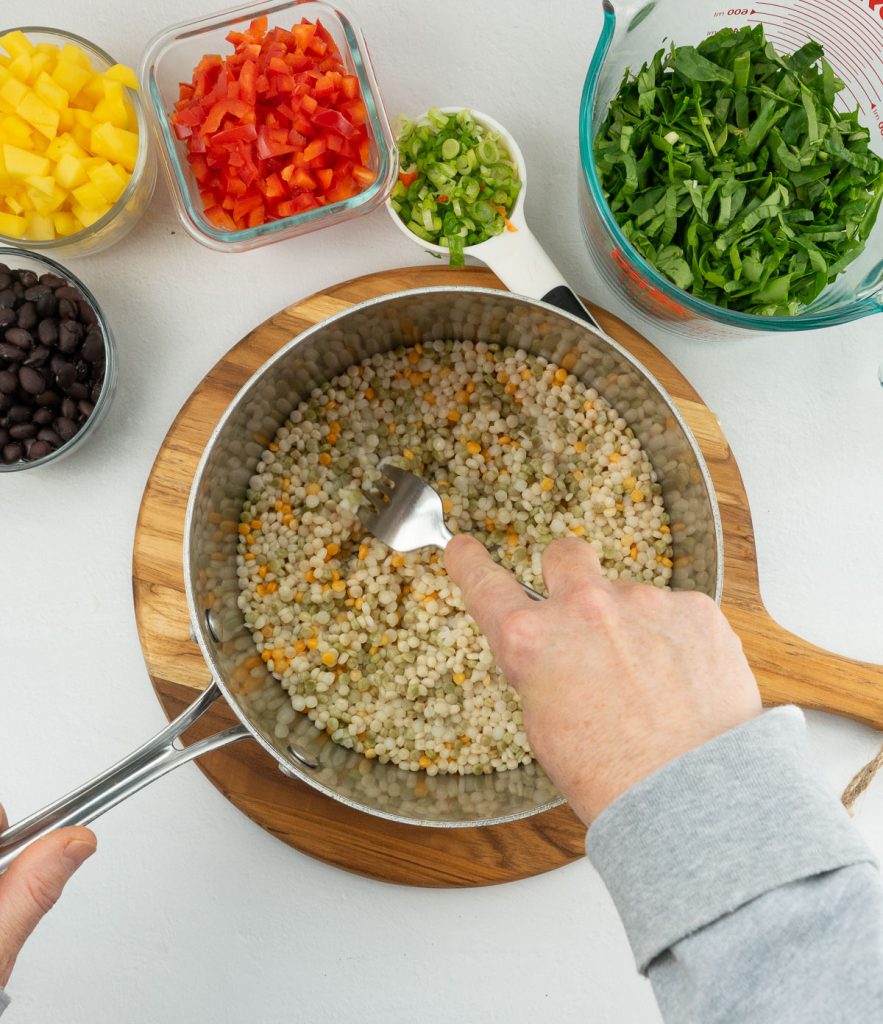 pan of couscous, hand fluffing couscous with a fork, other ingredients in small bowl next to pan