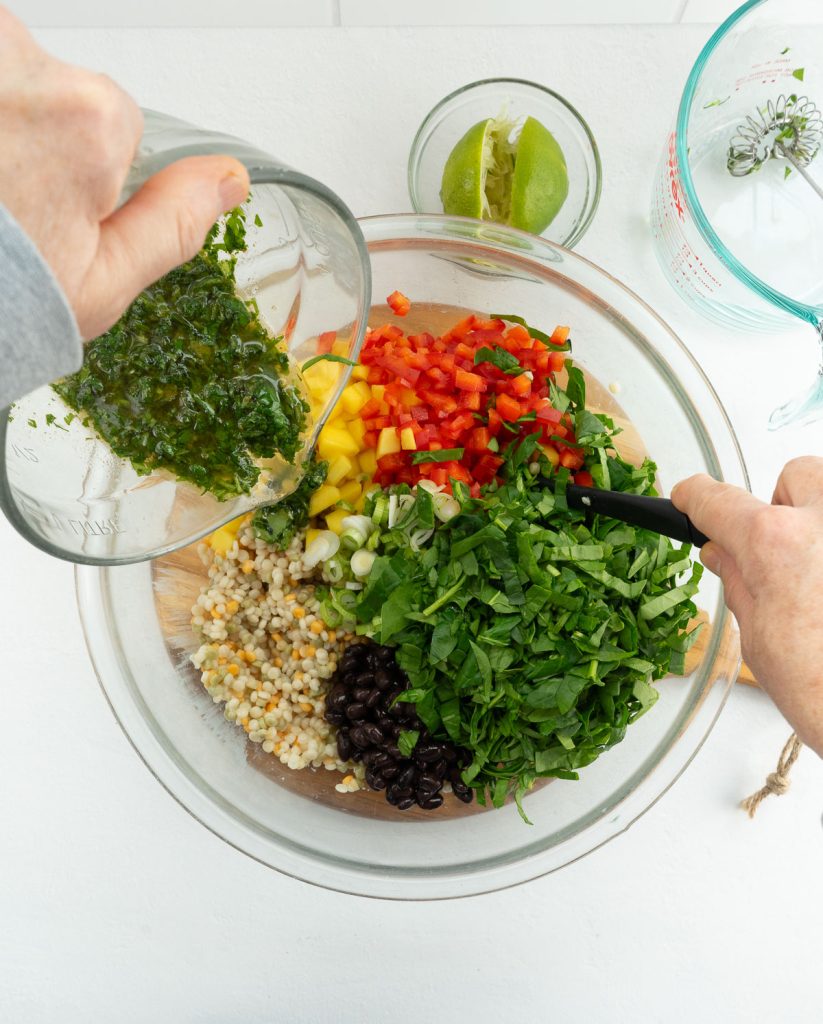 hand pouring dressing over salad ingredients. salad is in a glass bowl on a gray counter top. 