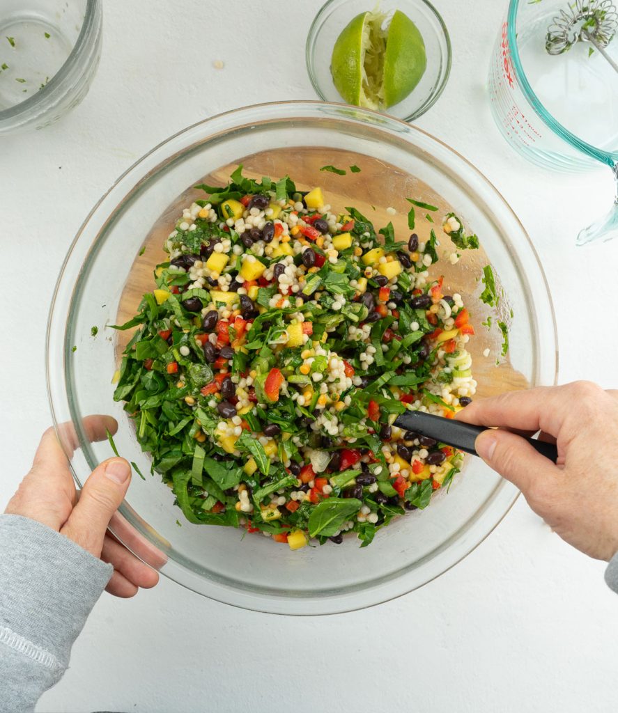 mixing mediterranean couscous sald in a glass bowl on a gray countertop limes in a small glass bowl at the top 