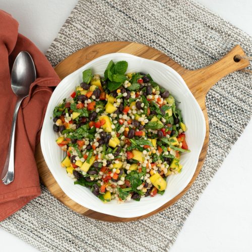 white bowl of mediterranean couscous salad on a wood cutting board with a tan place mat and a silver spoon on a rust colored napkin