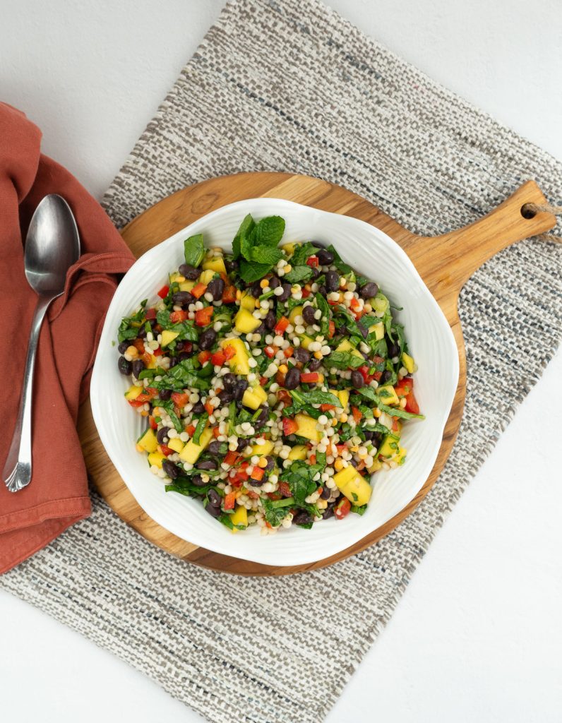 white bowl of mediterranean couscous salad on a wood cutting board with a tan place mat and a silver spoon on a rust colored napkin