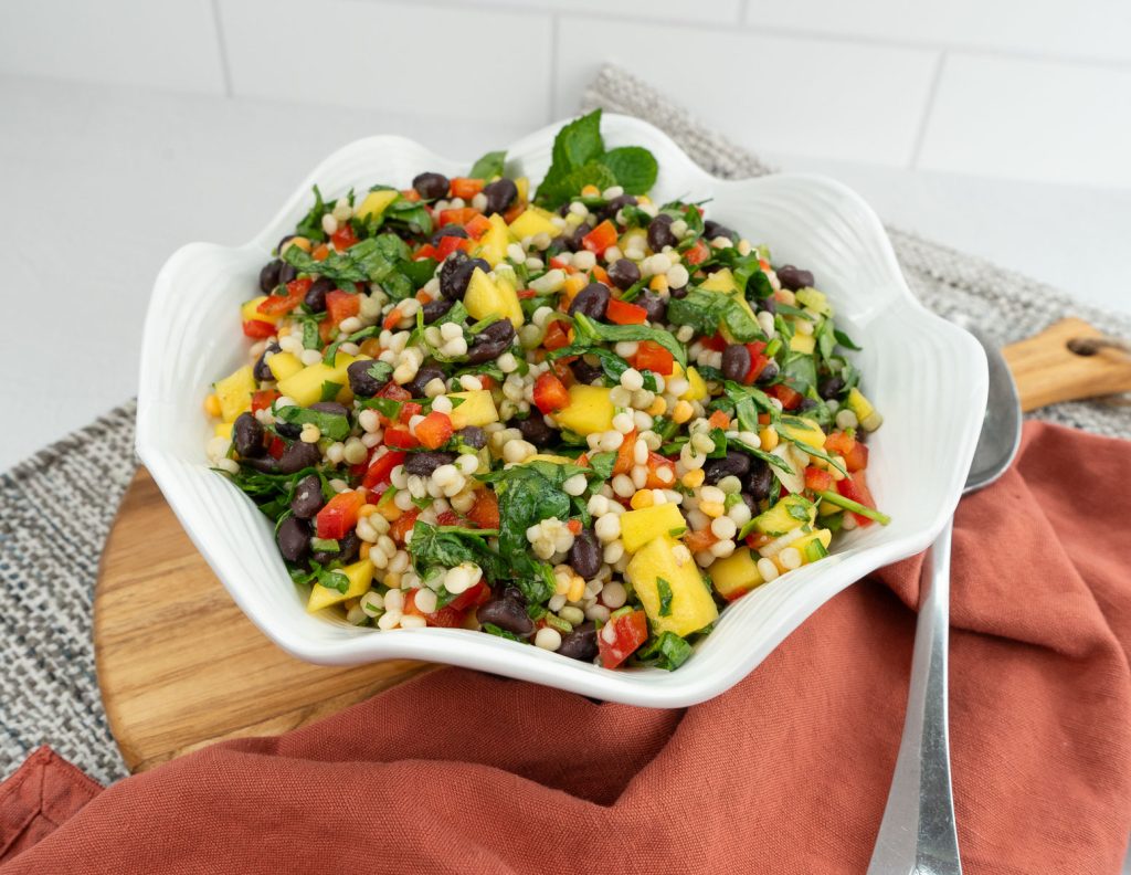 Mediterranean Couscous Salad in a white bowl placed on a round wood board with a silver spoon to the side and rush colored cloth in foreground