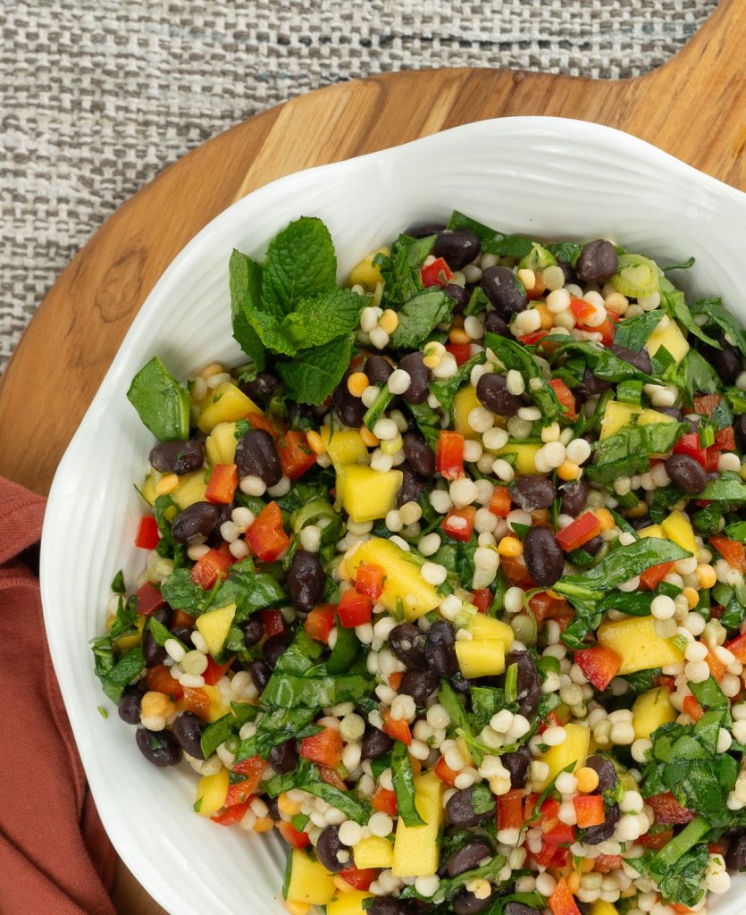 close up of mediterranean couscous salad in a white bowl on a wood board with tan placemat in background