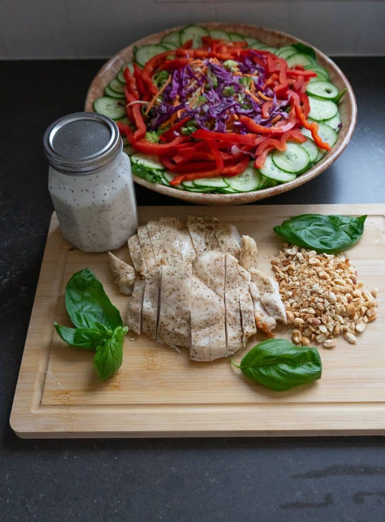 Bowl of prepared salad, sliced chicken, chopped peanuts, basil leaves and a jar of salad dressing on a wood cutting board in front of the bowl of salad