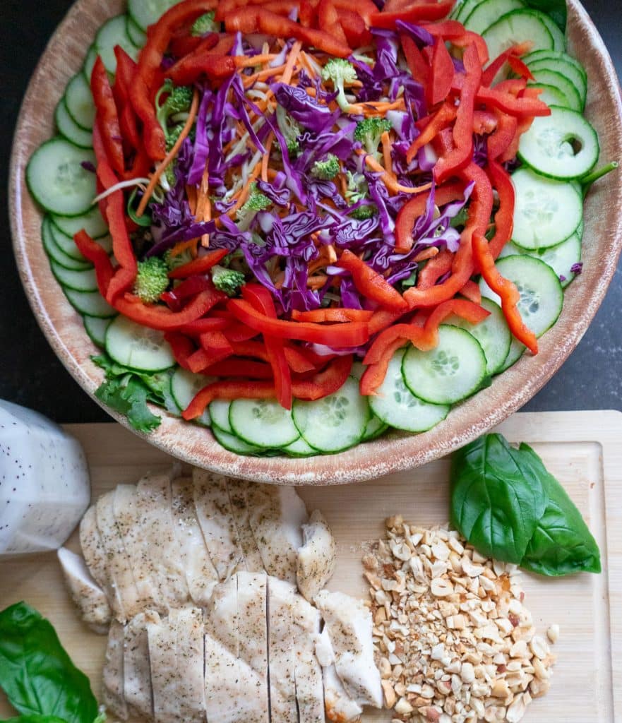 Bowl of prepared salad, sliced chicken, chopped peanuts, basil leaves and a jar of salad dressing on a wood cutting board in front of the bowl of salad