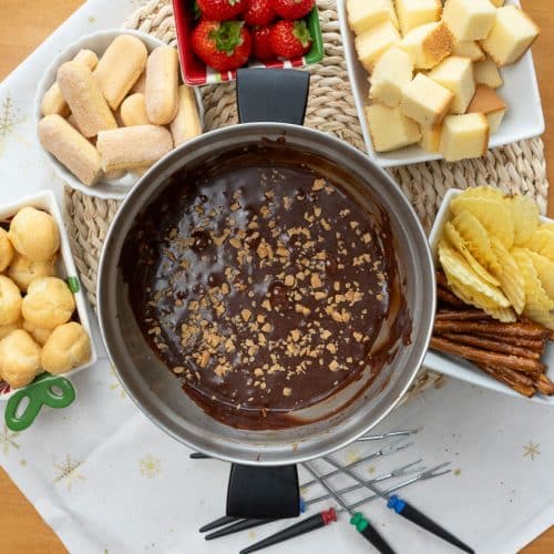 top view of a fondue pot with chocolate fondue in it with toffee bits sprinkled on top fondue pot is surrounded with food to dip in the chocolate