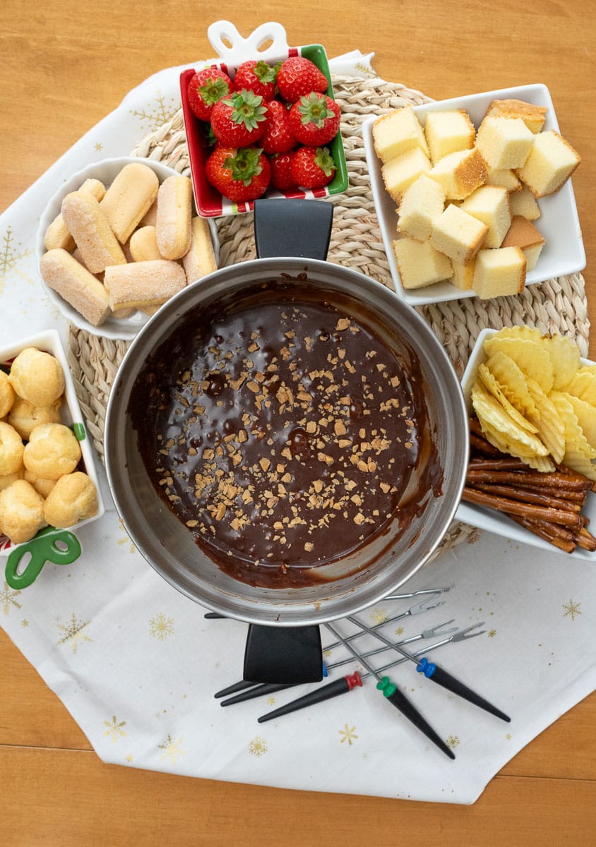 top view of a fondue pot with chocolate fondue in it with toffee bits sprinkled on top fondue pot is surrounded with food to dip in the chocolate