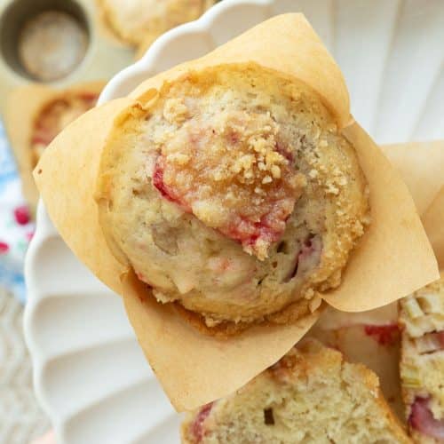 strawberry rhubarb muffin in a brown paper muffin paper liner on a plate with another muffin cut in half held over a table with the muffin pan full of baked muffins