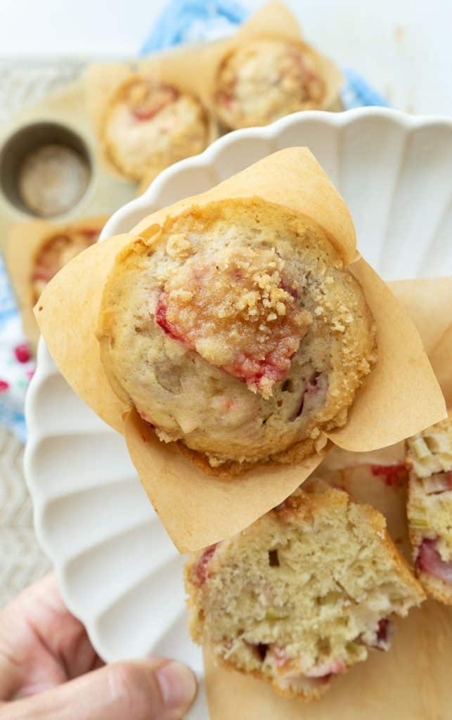 strawberry rhubarb muffin in a brown paper muffin paper liner on a plate with another muffin cut in half held over a table with the muffin pan full of baked muffins