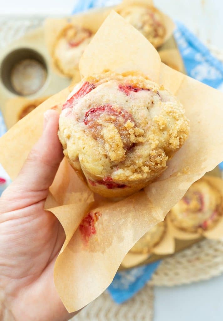 strawberry rhubarb muffin in a brown paper muffin paper liner on a plate with another muffin cut in half held over a table with the muffin pan full of baked muffins