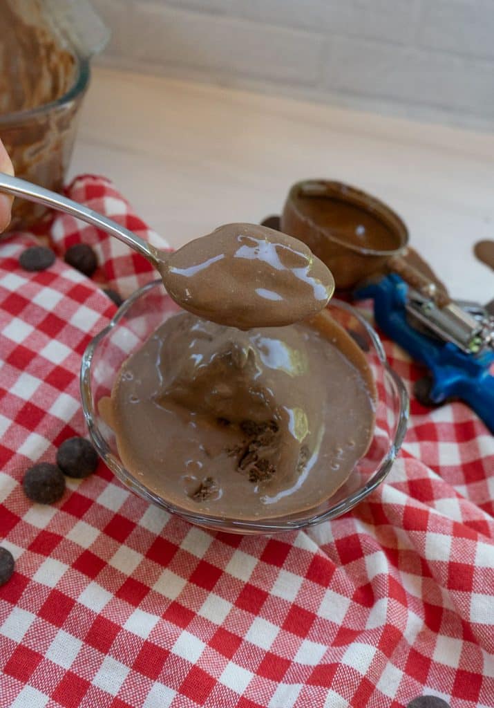 photo of brownie batter ice cream in a glass dish on a red and white gingham cloth with the ice cream scoop dripping on the table and chocolate chips on the table a spoon has ice cream dripping of it over the bowl of ice cream