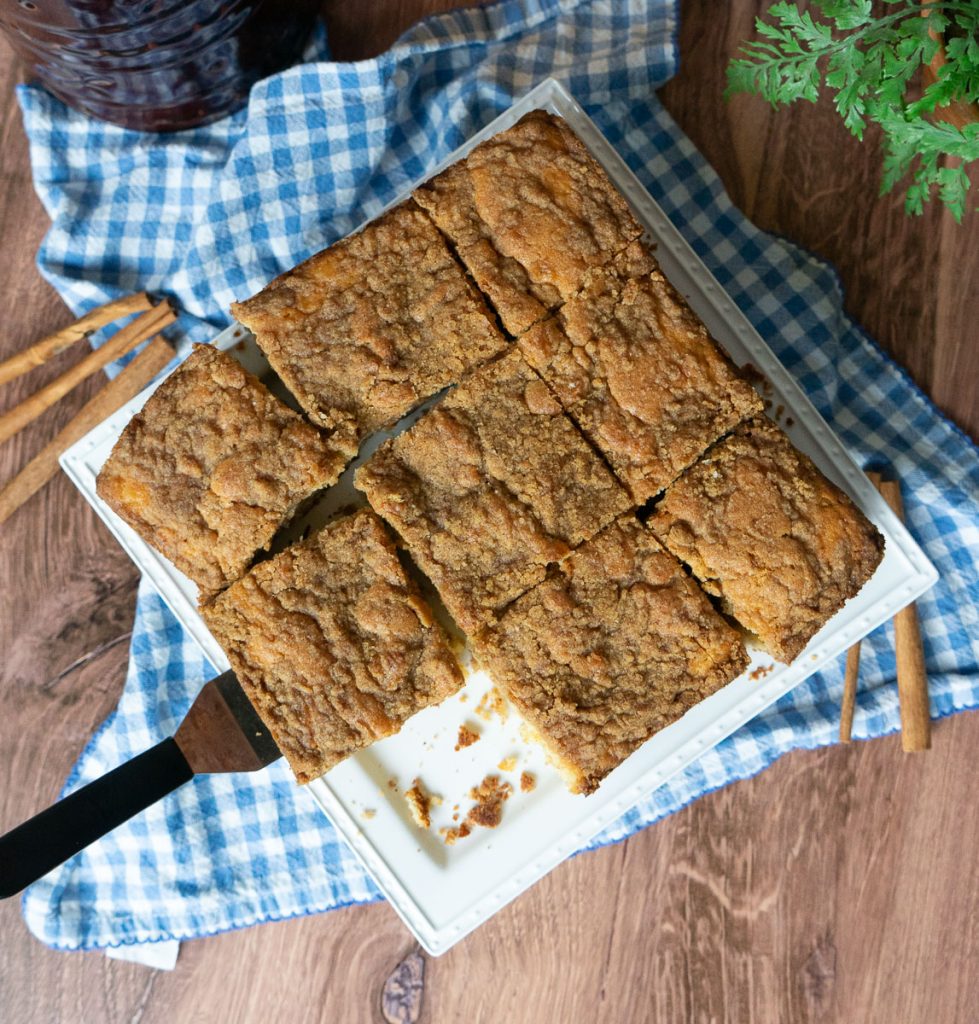top view of coffee cake cut into 9 pieces with one piece missing and a spatual under another piece ready to serve. cake plate is on a blue and white cloth